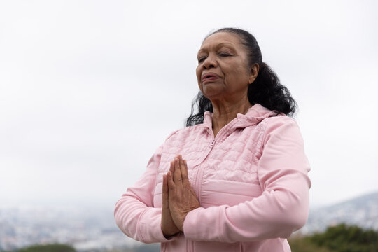 Senior African American woman standing on hill closing eyes holding prayer pose wearing pink jacket