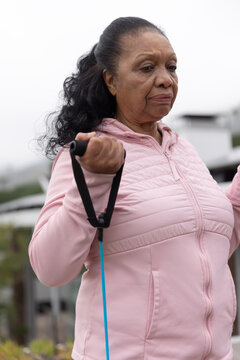 Senior African American woman exercising in park in pink zip-up jacket holding blue resistance band