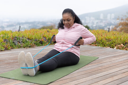 Senior African American woman in pink jacket black leggings exercising on green mat using blue band