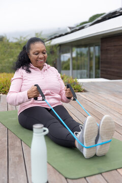 Senior African American woman in pink hoodie rowing on green mat on deck using band, bottle