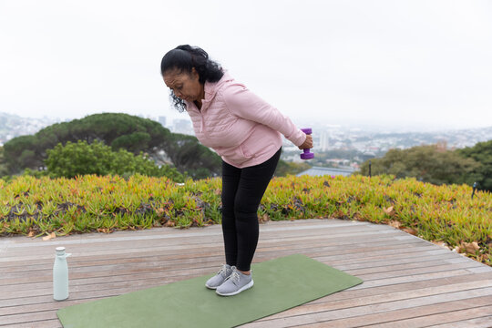 Senior African American woman bending on green mat on deck in pink jacket holding purple dumbbell