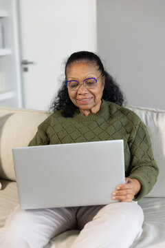 Senior African American woman sitting on sofa at home holding silver laptop wearing olive sweater