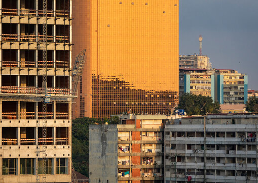 Golden glass skyscraper reflects the sunset beside an unfinished concrete tower and aging apartment blocks, Luanda Province, Luanda, Angola