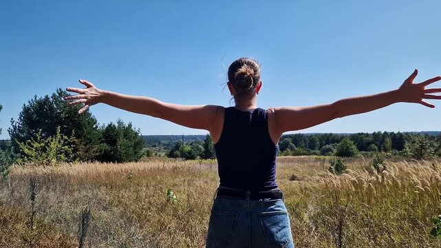 Woman with outstretched arms looking at the forest and field under the bright blue sky. Deep inhale and exhale, breathing techniques for relaxation, connection with nature, summer walk, early autumn
