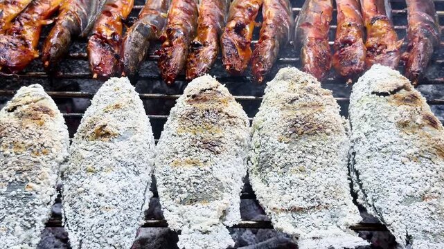 Grilled catfish on skewers and salt-crusted tilapia cooking over hot charcoal at an Asian street food market stall. Fresh barbecue seafood meal prepared outdoors on open fire.