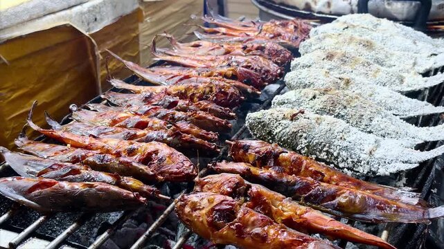 Grilled catfish on skewers and salt-crusted tilapia cooking over hot charcoal at an Asian street food market stall. Fresh barbecue seafood meal prepared outdoors on open fire.