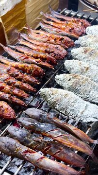 Grilled catfish on skewers and salt-crusted tilapia cooking over hot charcoal at an Asian street food market stall. Fresh barbecue seafood meal prepared outdoors on open fire.