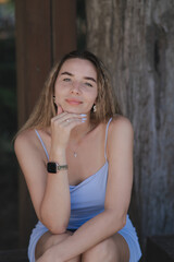 Fototapeta premium A beautiful young woman in a light blue dress with thin straps sits outdoors, resting her chin on her hand, and smiles gently.