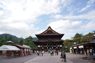 Obraz premium 日本の古都にある荘厳な寺院の境内が広がる夏の風景。中央には伝統的な木造建築の本堂が堂々と建ち、多くの参拝客や観光客が石畳の広場を散策している。背後には緑豊かな山々が連なり、穏やかな雲が浮かぶ青空が広がる長野県善光寺の一日。