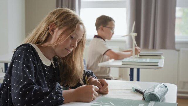 Caucasian schoolgirl writing in notebook during science class while classmate studies wind turbine model. Clip suitable for STEM curriculum promo and green energy education campaigns