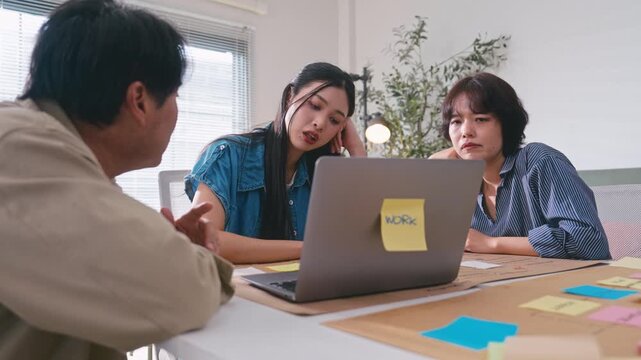 Professional Asian business team gathering around desk to analyze data on laptop screen.