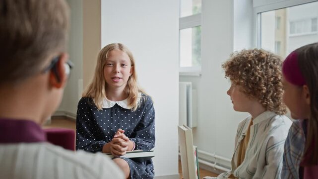 Caucasian schoolgirl speaking during group discussion with classmates in classroom, over shoulder view. Footage suits elementary education campaigns, reading clubs, and school ads
