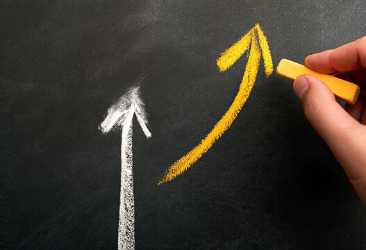 Hand draws a yellow upward arrow overtaking a white upward arrow on blackboard