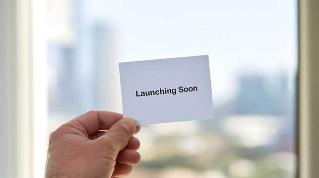 A person hold a white paper card with launching soon text on it in front of a bright window with city view for business promotion concept