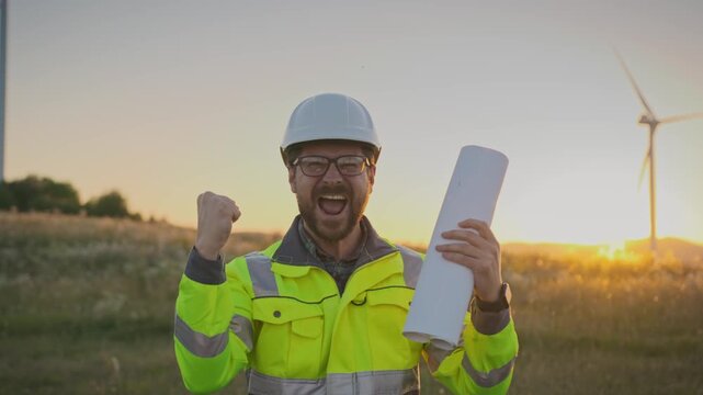 Smiling Caucasian male engineer in helmet and glasses wearing reflective jacket outdoors near turbines. Raising fist and celebrating success while standing in field during sunset after completing task