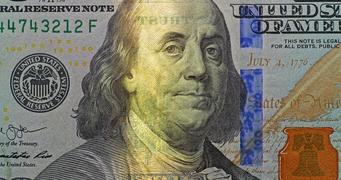 US one hundred dollar notes features Benjamin Franklin portraits and shifting golden reflections. This currency detail holds steady while light glides across printed security elements.