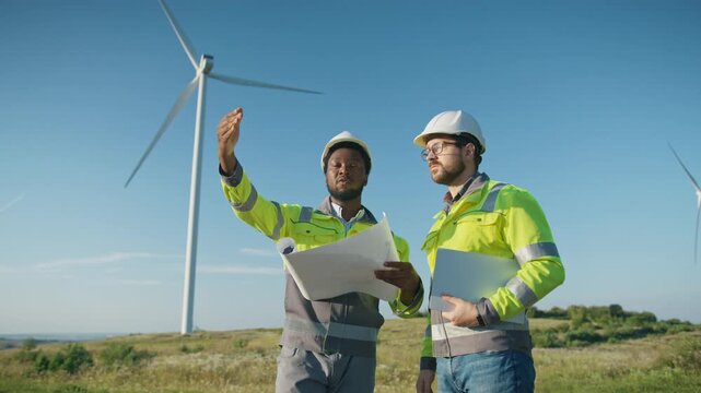 Focused African American male engineer wearing helmet and safety jacket holding print outdoors. Standing with colleague in glasses near wind turbine. Discussing project and pointing direction.