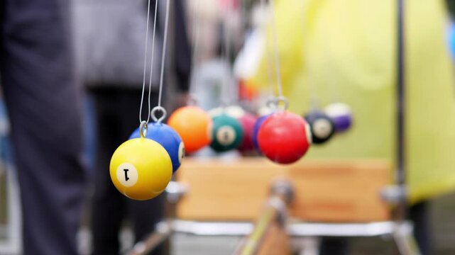 Colorful billiard balls hang on thin strings inside indoor game room, with yellow ball in sharp focus and red, blue, orange, purple, teal, and black balls softly blurred behind, under warm light