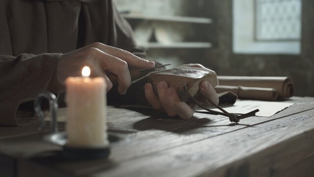 Hands of a medieval monk sharpening a penknife blade on a whetstone at a wooden table near a burning candle inside a scriptorium