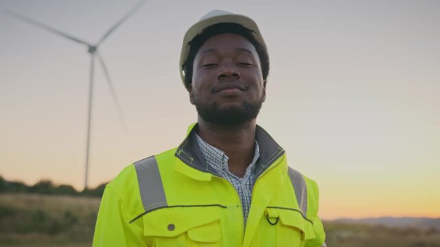 Focused African American male engineer wearing reflective jacket adjusting helmet outdoors. Standing near wind turbine in field preparing for work on renewable energy project during sunset.