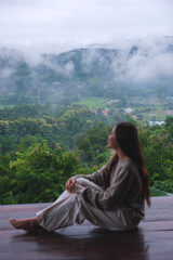 Obraz premium Vertical image of a woman sitting on wooden balcony with a beautiful mountain views on cloudy morning