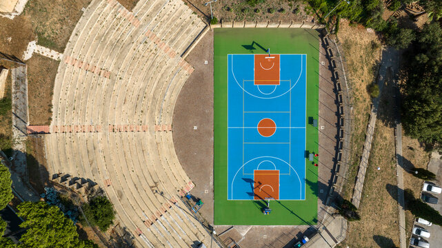 Aerial zenithal view of an empty outdoor basketball court with a blue playing surface, next to the curved concrete steps of a small grandstand, seen from above on a sunny day. The park is empty.