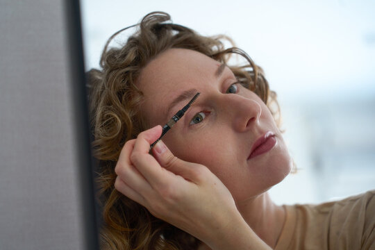 Woman grooming her eyebrows with a spoolie brush in front of a mirror.
