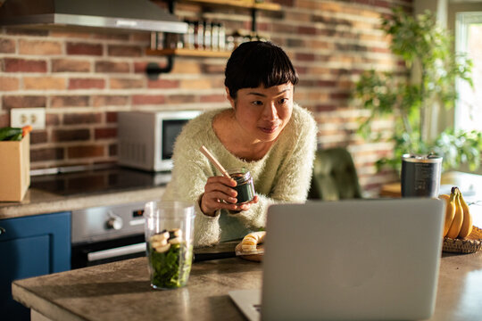 Woman preparing green smoothie while watching laptop in home kitchen