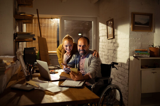 Smiling couple using smartphone and laptop at home office, man in wheelchair