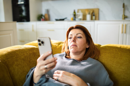 Middle-aged woman relaxing on sofa using smartphone in modern home kitchen