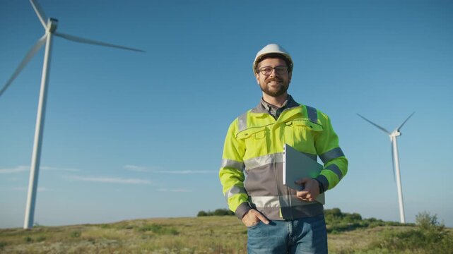 Smiling Caucasian male engineer with beard and glasses wearing helmet and reflective jacket standing near wind turbines. Holding laptop and inspecting energy field. Considering technical solutions.