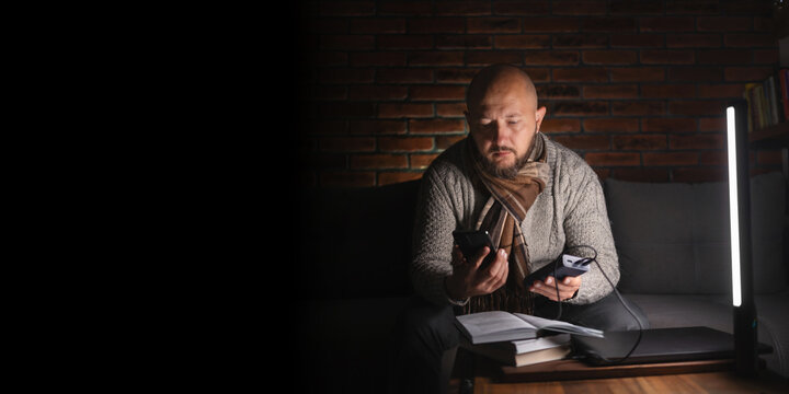 Ukrainian man on sofa in warm clothes using smartphone and holding power bank near led lamp, laptop and books on table during power outage in cold dark home caused by Russian attacks on energy grid