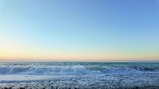 Storm waves wash ashore on a pebble beach under a soft pastel sunset sky.