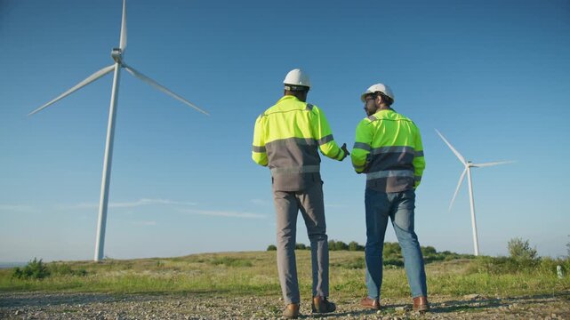 Two male engineers wearing helmets and reflective jackets while walking together outdoors near wind turbines. Carrying documents and moving across field. Monitoring renewable energy project progress.