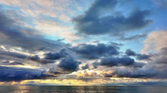 Dramatic clouds illuminated by golden sunset light stretch across the sky above a calm ocean horizon. Timelapse.