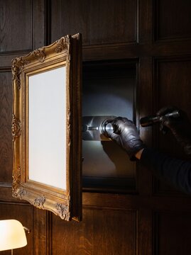 Gloved hands cracking hidden wall safe behind gold frame