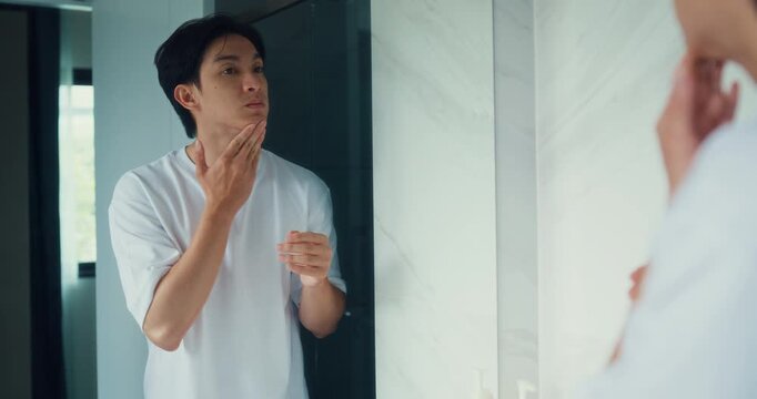 Young Asian man applies moisturizer cream to face while looking in bathroom mirror at home. Daily skincare routine, facial hydration, healthy complexion, beauty care and cosmetic wellness concept.