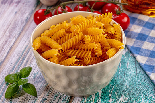 Uncooked fusilli pasta in ceramic bowl with fresh basil, cherry tomatoes and olive oil on rustic wooden table with blue checkered cloth