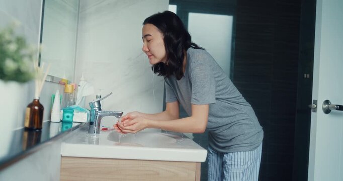 Young Asian woman washes face with cleansing foam in bathroom mirror at home. Daily skincare beauty facial routine, Healthy fresh moisture cosmetic lotion, Personal hygiene wellness care concept.