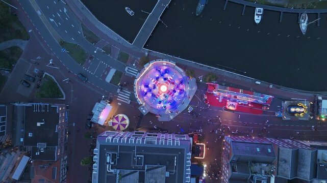 Top down aerial drone footage of a colorful fairground at dusk with a large spinning amusement ride, bright lights, visitors and attractions beside the waterfront.