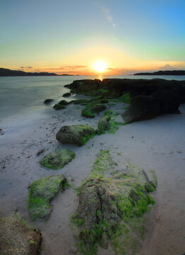 Golden sunset over a tropical beach with green moss-covered coastal rocks at Gili Kedis, Sekotong, West Lombok, Indonesia.