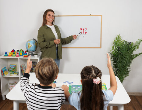 Boy and girl raise their hands to answer a female teacher pointing at an addition problem on a whiteboard during an interactive elementary mathematics lesson in classroom.