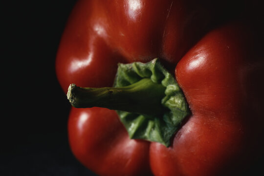 Macro shot of ripe red bell pepper with vibrant green stem in moody lighting on dark background. Fresh raw vegetable for organic recipes, culinary menus, nutrition, and healthy eating.