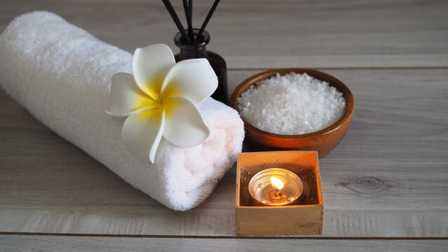 Spa wellness arrangement featuring a rolled white towel decorated with a white plumeria flower, a lit tealight candle in a wooden box, a wooden bowl filled with white bath salts, and a reed diffuser b