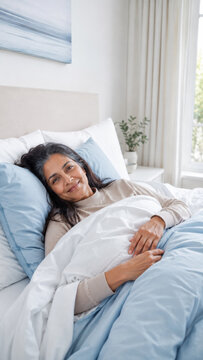 Middle-aged woman smiles while resting under white bedding in a bright bedroom. Comfortable home setting with soft natural light and calm wellness concept.