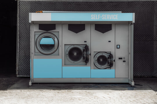 Street laundromat with two open washers, a dryer and blank blue signage panels for custom text, branding or service information on a metal self-service kiosk against a dark fence