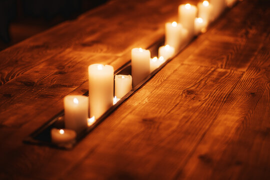 Ivory pillar candles of varying heights burn in a recessed row between textured wooden planks, casting warm amber light across a rustic tabletop with soft blur and an intimate evening mood