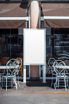 Empty white advertising board stands between outdoor bistro tables and ornate metal chairs, offering a vertical template for menus or promotions against glass windows and brown awnings