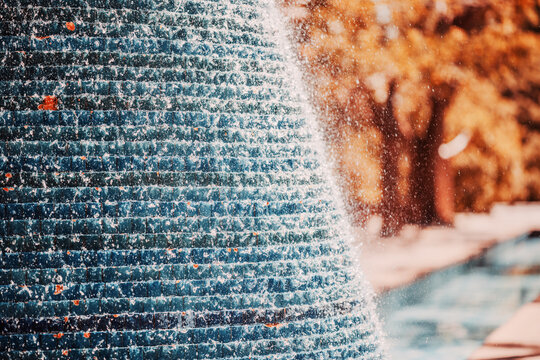 Selective focus captures sparkling spray cascading over blue fountain tiles in the foreground, while orange foliage and a pool blur behind, contrasting cool water with warm autumn colors