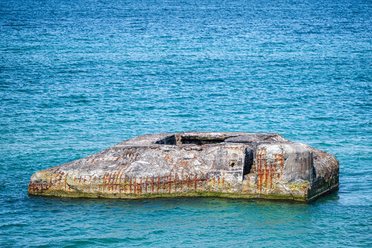 An old concrete military bunker partially submerged in clear sea water at Vigso beach, showing a rusty entrance and weathered walls with green algae.